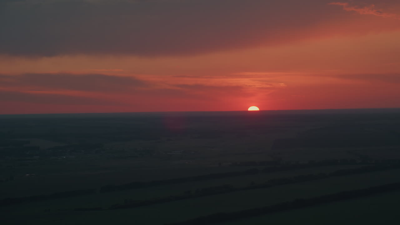 Vibrant red orange sky with dark cloud layers at dusk as sun half dips below farmland horizon casting subtle lens flare over expansive rural landscape in serene evening glow