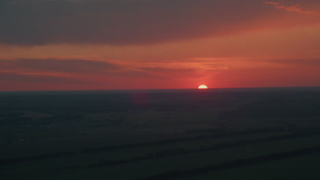 vibrant red orange sky with dark cloud layers at dusk as sun half dips below farmland horizon casting subtle lens flare over expansive rural landscape in serene evening glow