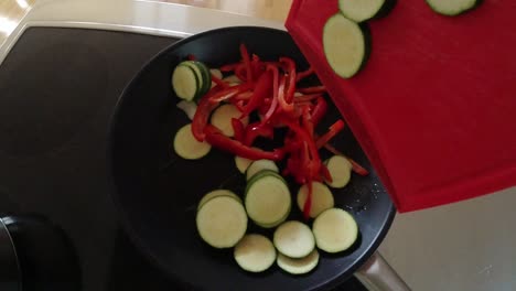 Slow-motion video of a chef dropping vegetables into the frying pan in a non-commercial kitchen. From the top down perspective capsicum and zucchini slices for stir fry.