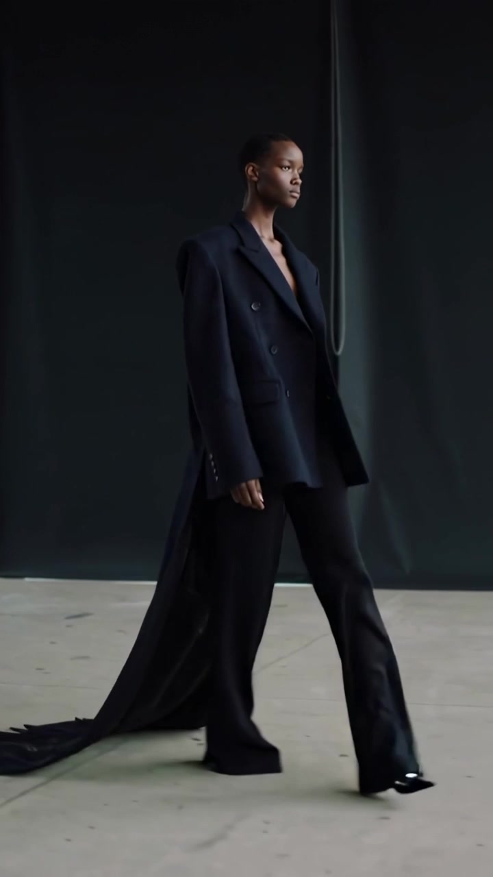 Vídeo vertical: Modelo andando a meio passo vestindo blazer preto com painel e calças largas em passarela de estúdio
