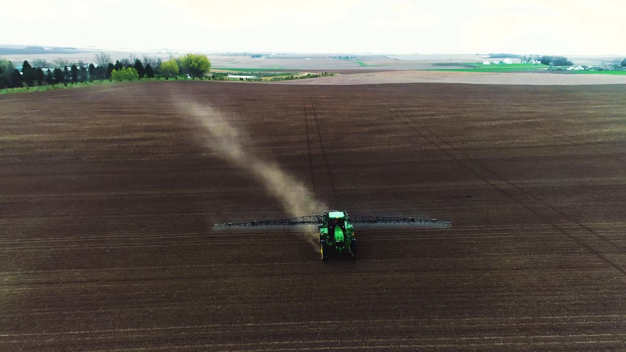 Tomada de drone subindo de um grande pulverizador John Deere atravessando solo preto para revelar a fazenda ao fundo