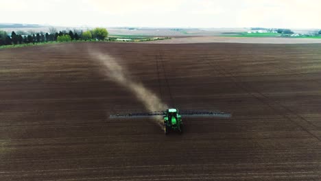 Pan-up-drone-shot-of-large-John-Deere-sprayer-driving-across-black-soil-to-reveal-farm-in-the-background