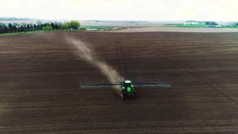 Toma-Panorámica-De-Un-Dron-De-Un-Gran-Rociador-John-Deere-Conduciendo-A-Través-Del-Suelo-Negro-Para-Revelar-La-Granja-En-El-Fondo