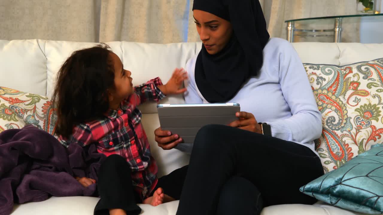 Young mother using a digital tablet with her daughter on the sofa at home 4k