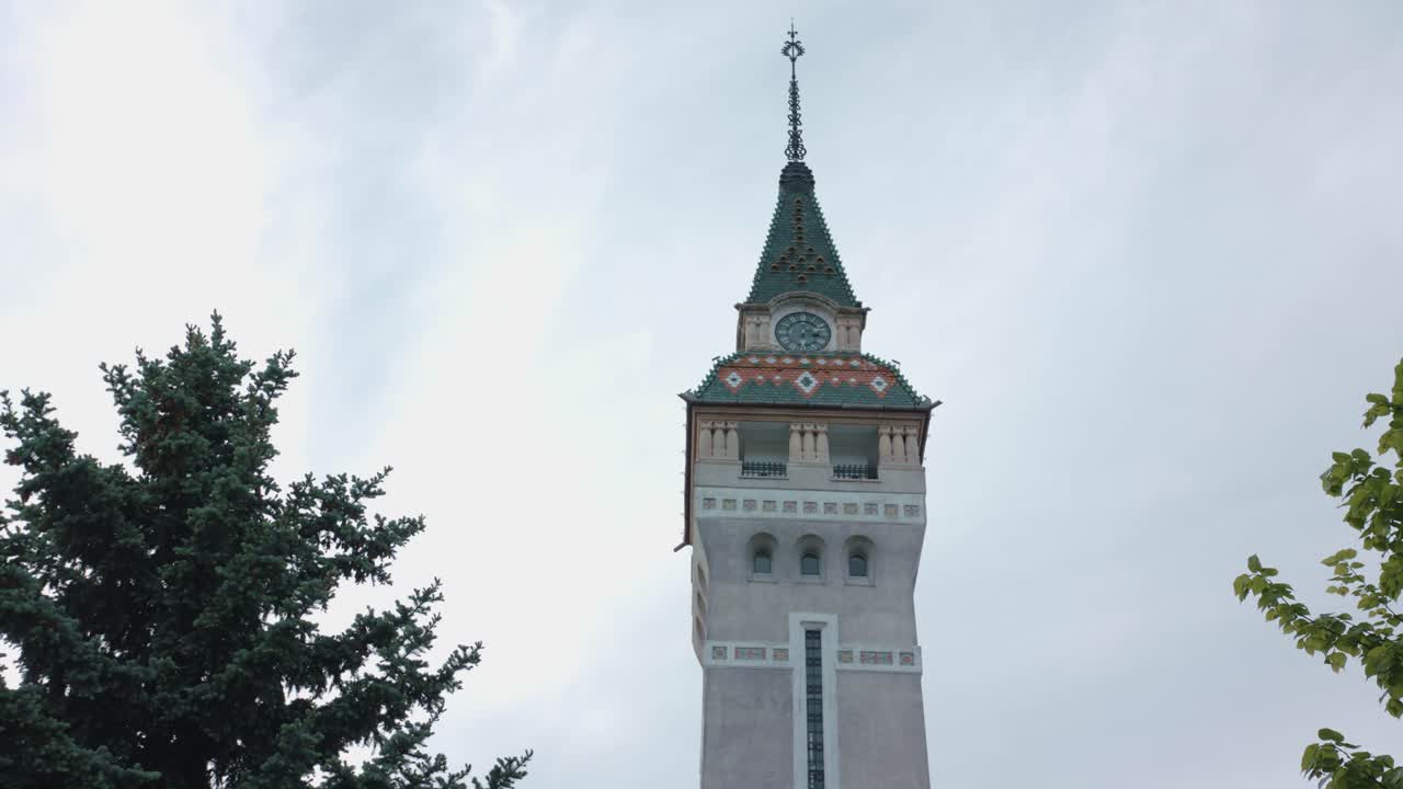 A low-angle, slow-moving shot of the historic clock tower in Targu Mures. The ornate spire is framed by green trees, standing tall against a dramatic, grey, overcast sky