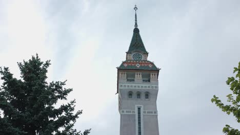 a low-angle, slow-moving shot of the historic clock tower in targu mures. the ornate spire is framed by green trees, standing tall against a dramatic, grey, overcast sky