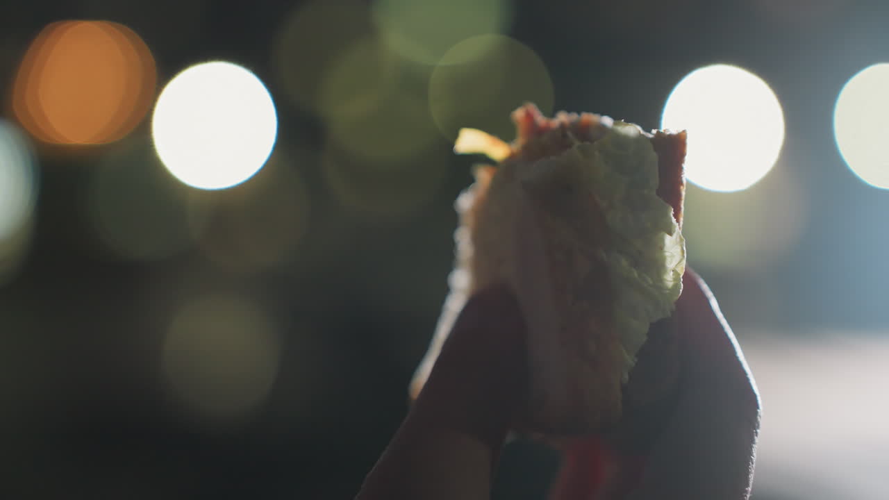 Close up hands of fair skinned woman holding crusty bread sandwich outdoors on bench at urban park twilight under glowing lights enjoying post workout snack moment with reflective calm mood