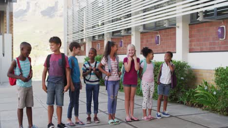 diverse group of schoolchildren wearing backpacks smiling and standing in a row at school yard