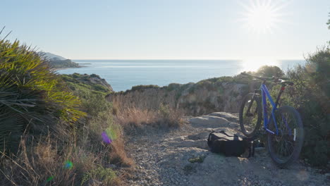 drone flies through coastal scene towards ocean as mountain bike is parked with backpack beside it in foreground during morning light