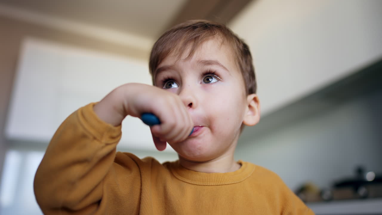 Adorable baby boy takes a full spoon into his mouth. Kid chews his food and waves his spoon in front of camera. Low angle view.