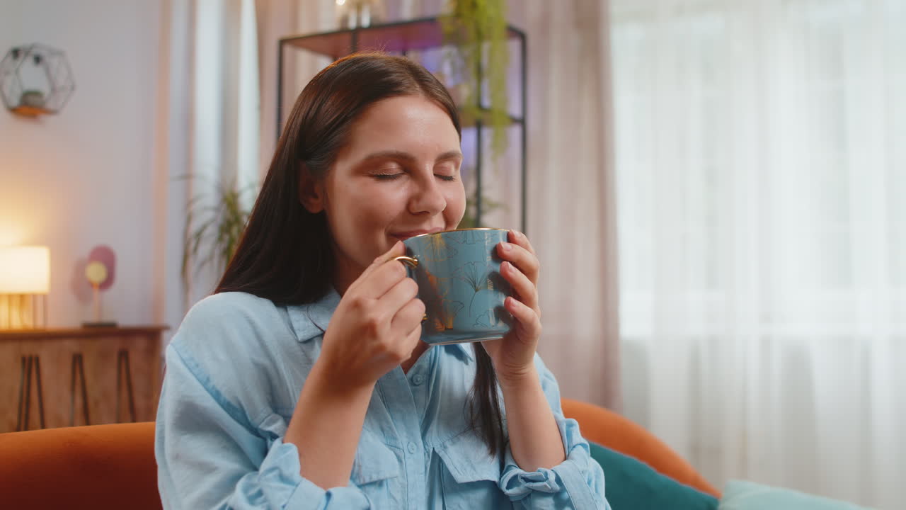 Ragazza giovane attraente e sorridente che beve una tazza di caffè o tè alle erbe seduta sul divano di casa
