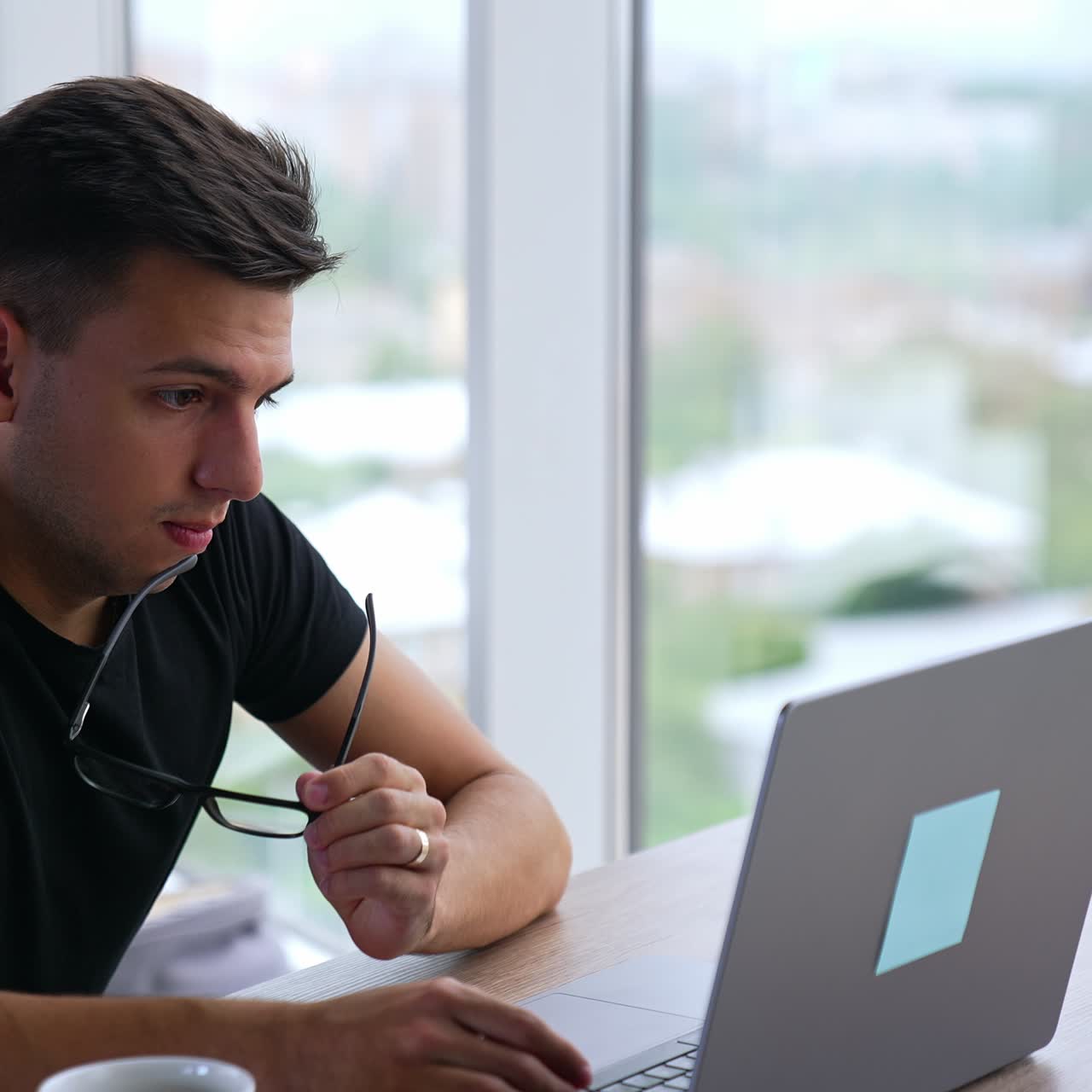 Busy young man in front of laptop puts off glasses and looks thoughtfully on the screen. Office backdrop indoors