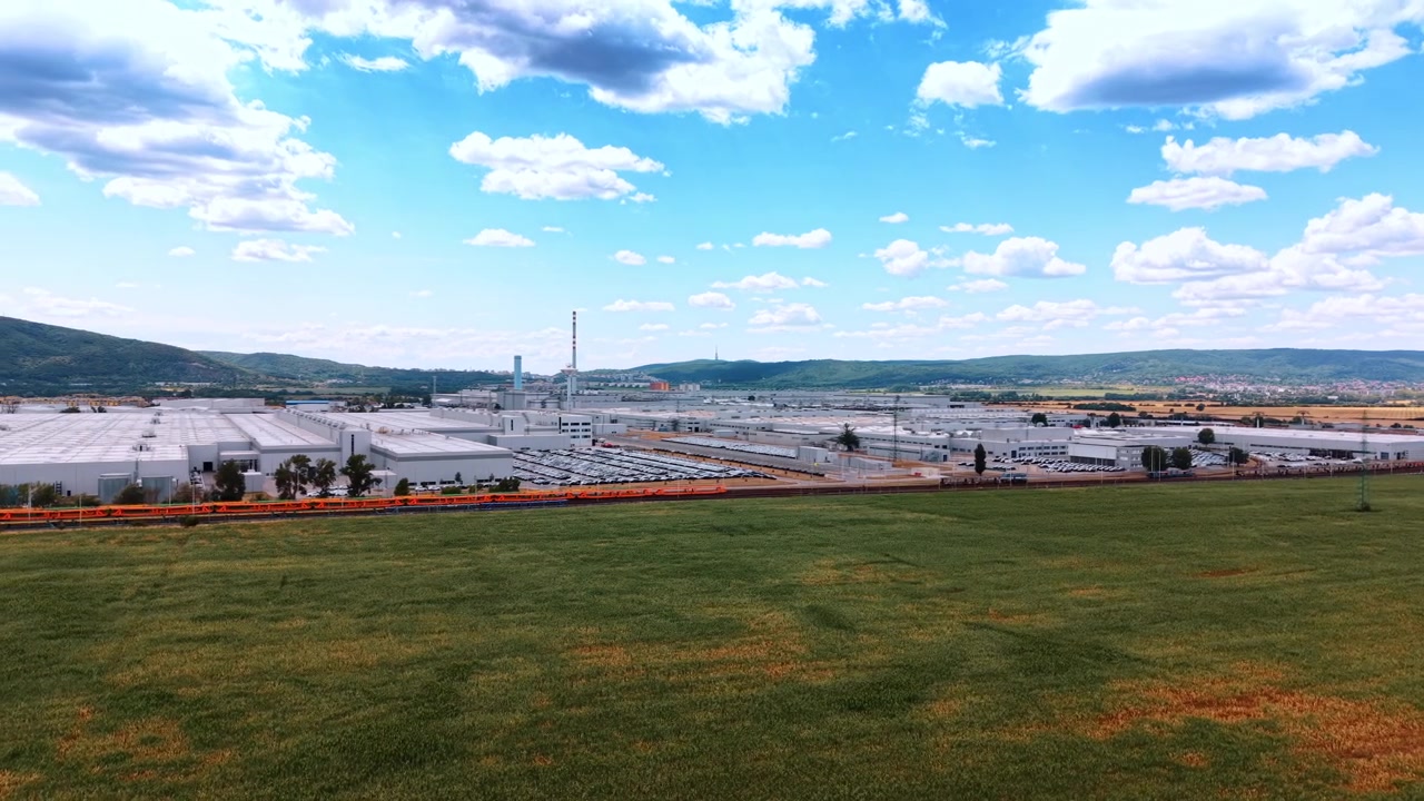 Automobielfabriek en spoorlijn gezien vanuit een lage luchthoek. Een goederentrein passeert een uitgestrekte parkeerplaats naast fabrieksgebouwen, omlijst door groen gras en verspreide wolken.