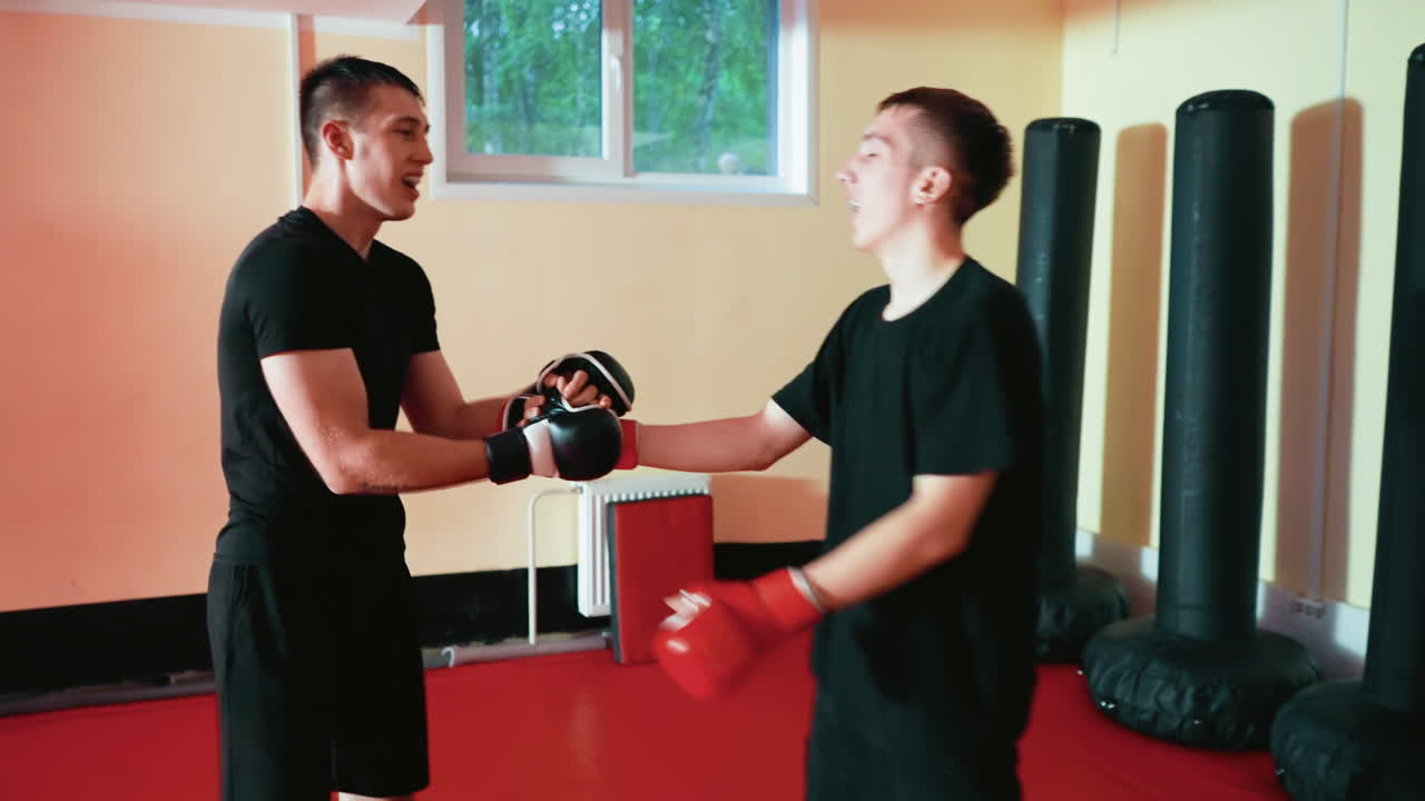Wrestlers engaged in intense sparring session grapple in close stance wearing gloves on red mat gym floor,hug and shake each other,with punching bags in background