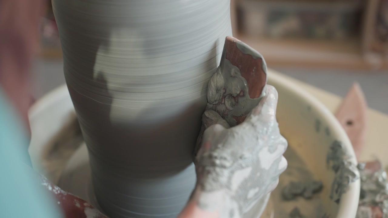Clay sculpture under gentle touch. Close view of hands sculpting ceramic with sponge smoothing rim. Detailed shot of artisan hands shaping moist clay cylinder on spinning wheel using sponge