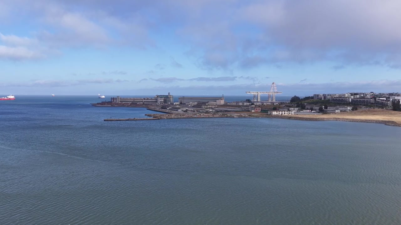 Il filmato aereo cattura un vasto porto costiero, un ampio porto, gru industriali, una costa sabbiosa e acque blu calme sotto nuvole sparse. La luminosa luce del giorno illumina la scena panoramica sul mare.