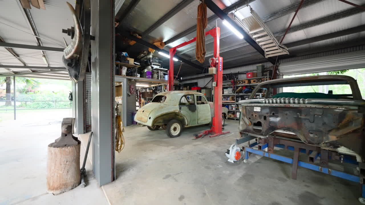 Ripresa in allontanamento da un'officina residenziale che restaura vecchie auto arrugginite e malandate, in un garage ben illuminato e fornito di attrezzi, con l'esterno visibile