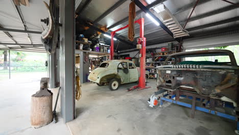 pullback from residential work shed restoring old beater rusted cars in garage well lit and stocked with tools outside