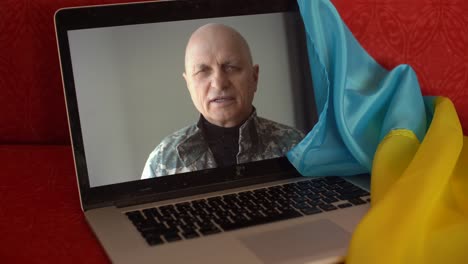 soldier in military uniform sits on sofa at home using webcam laptop for communication with family. ukrainian flag on the background. military actions in ukraine. free democratic people in the state