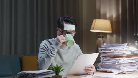 아픈 아시아인 남자가 집에서 문서와 함께 열심히 일하면서 물을 마시고 있습니다.