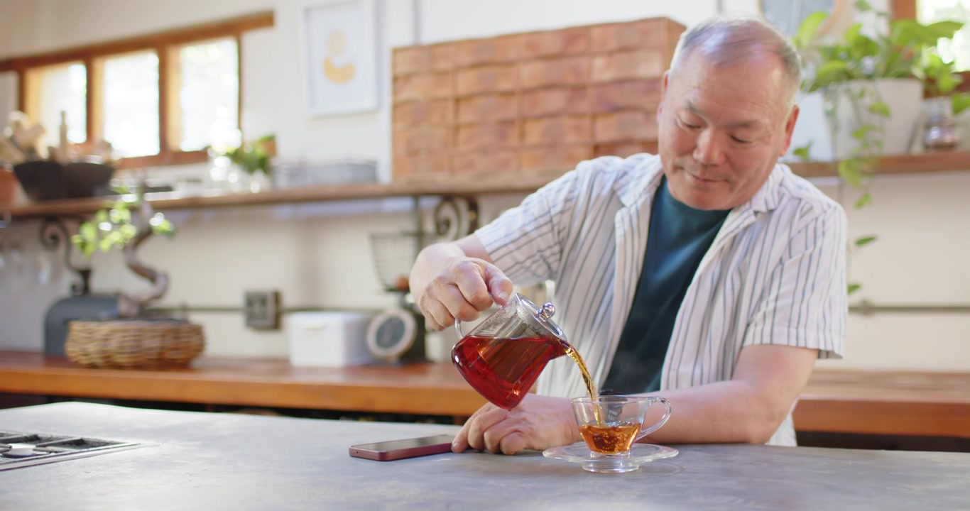 Anziano uomo asiatico che solleva una teiera, versando il tè sul bancone della cucina vicino a uno smartphone per riempire la tazza.
