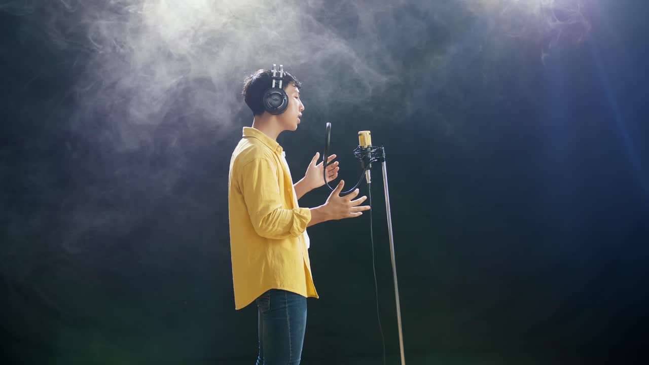 하 연기 검은 배경에 콘덴서 마이크로 노래하는 헤드폰을 가진 젊은 아시아 소년의 측면 시각