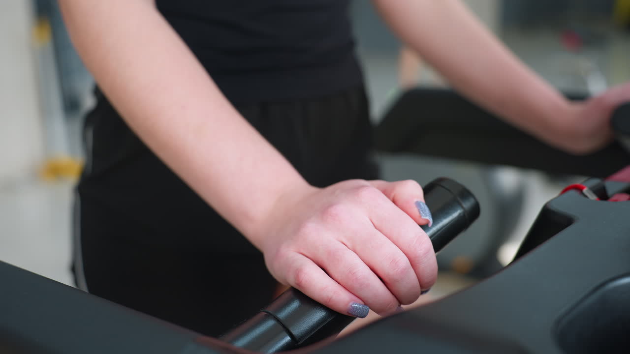 close up of young woman wearing black shorts gripping treadmill handle during gym workout, focus on hand with painted nails and blurred fitness environment in background