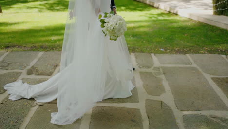 Una-Novia-Con-Un-Vestido-Blanco-Fluido-Y-Un-Novio-Caminando-Juntos-Por-Un-Camino-De-Piedra-En-Un-Jardín