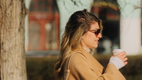 Pretty-Blonde-Elegant-Woman-in-Happy-Mood-Dressed-in-Beige-Coat,-Walking-Alone-in-Street-and-Drinking-Coffee,-Cinematic-Shot