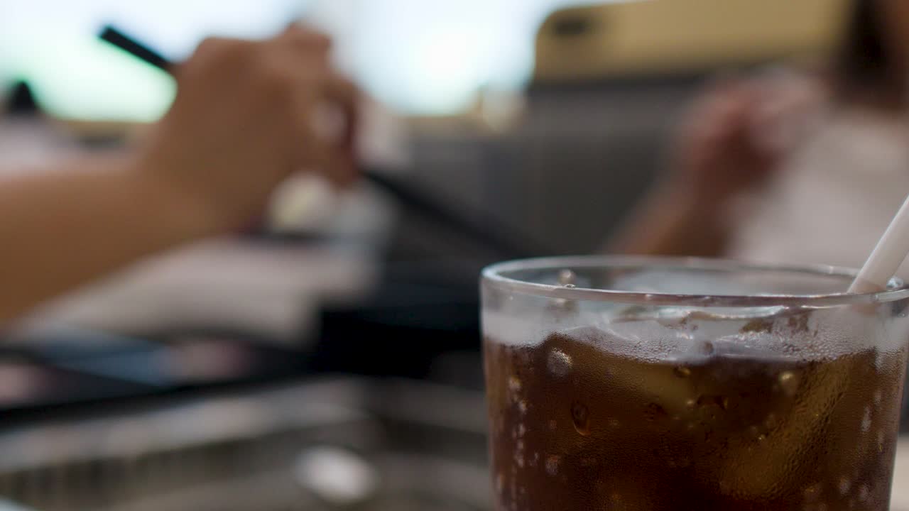 Primo piano di una cola fredda con ghiaccio e cannuccia in un ristorante affollato.