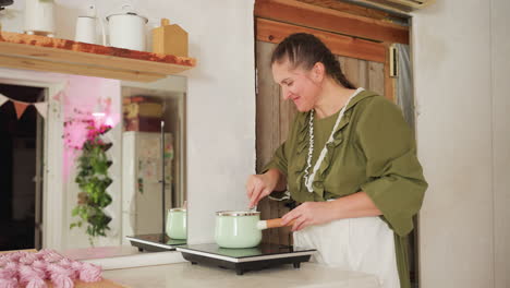 happy female cook in green dress prepares lunch using induction cooker while smiling in cozy kitchen with rustic wooden cabinets, large mirror reflecting background