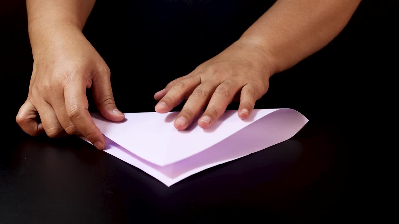 Person’s hands folding white paper on black table, step-by-step origami preparation, studio lighting