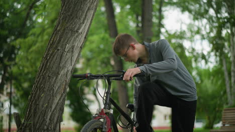 young boy wearing glasses holds the handlebar of parked bicycle in grassy field, adjusting the handlebar firmly while checking the brake functionality, he pauses to adjust his sneakers