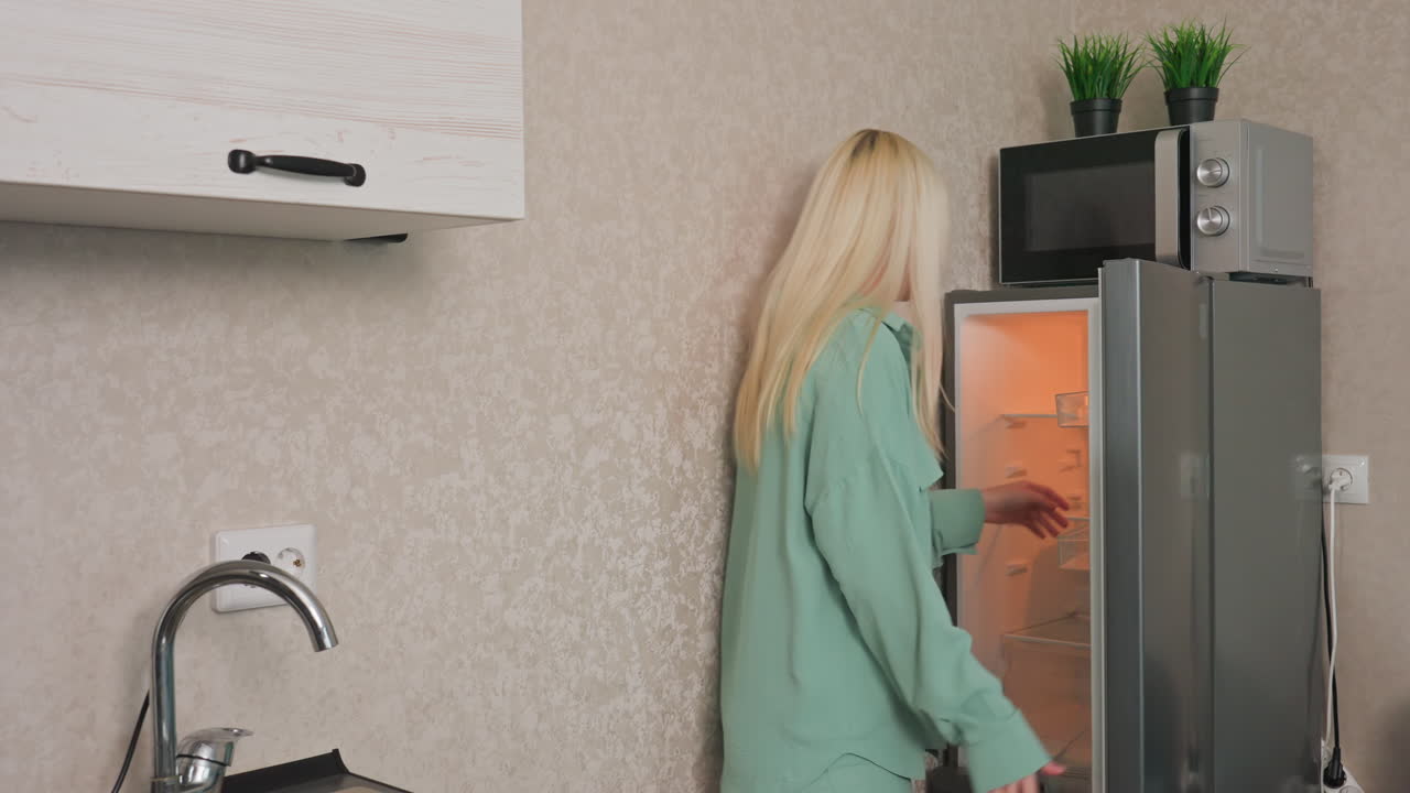 Two ladies in modern kitchen, one opening fridge, dropping item, handing it to friend near table with colorful bowls and teal couch, casual home moment with natural lighting