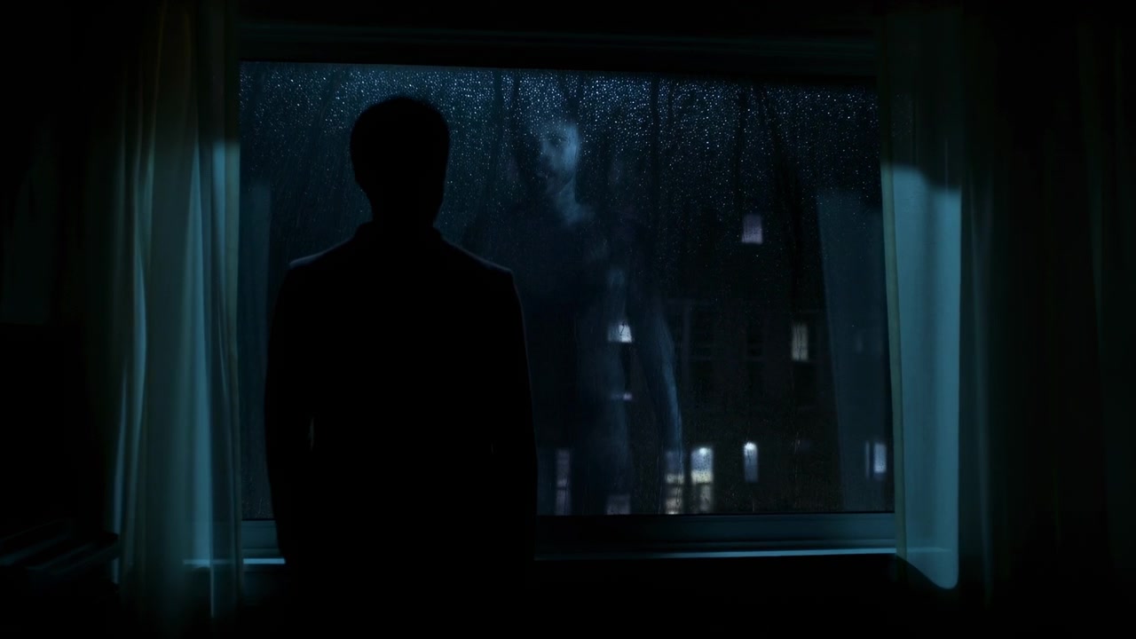 Una silhouette solitaria si staglia in un interno scuro accanto a una finestra coperta di gocce di pioggia, con tende che incorniciano la vista e deboli luci della città che brillano all'esterno