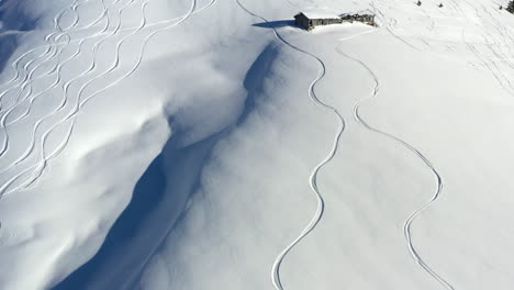 Aerial-shot,-lifting-up-to-show-fresh-ski-tracks-in-fresh-snow,-then-revealing-an-old-mountain-hut---barn-on-the-side-of-a-hill