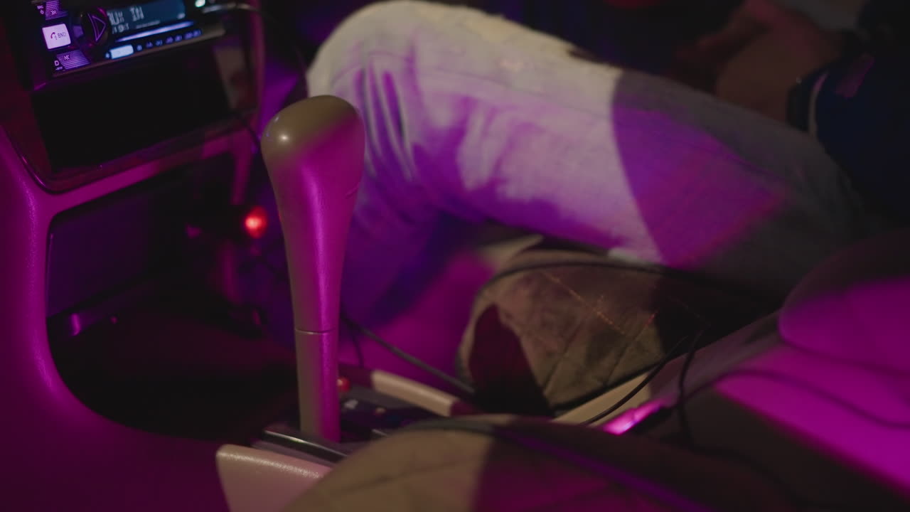 Interni notturni di un'auto, dettaglio della leva del cambio, persona tesa che cerca di ingranare la marcia sotto una luce ambientale viola, mano con giacca di jeans che esita vicino alla console, bagliore del cruscotto, flash dell'orologio da polso, atmosfera da passaggio in rideshare, movimento cauto