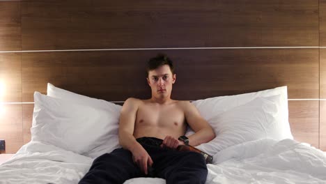 white caucasian male flipping channels in a hotel room on his bed in disdain with a bottle of whisky in his hand