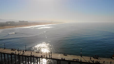 Wide,-sunrise-aerial-4k-drone-pullback-from-Huntington-Beach-to-reveal-pier---Diner-on-Pacific-Ocean-in-Southern-California,-early-morning-w--waves,-surfers,-fisherman,-shimmering-seas-visible