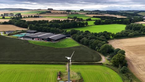 drone glides above green fields, wind turbine, and farm buildings under cloudy daylight, revealing rural landscape and sustainable energy infrastructure with smooth camera movement