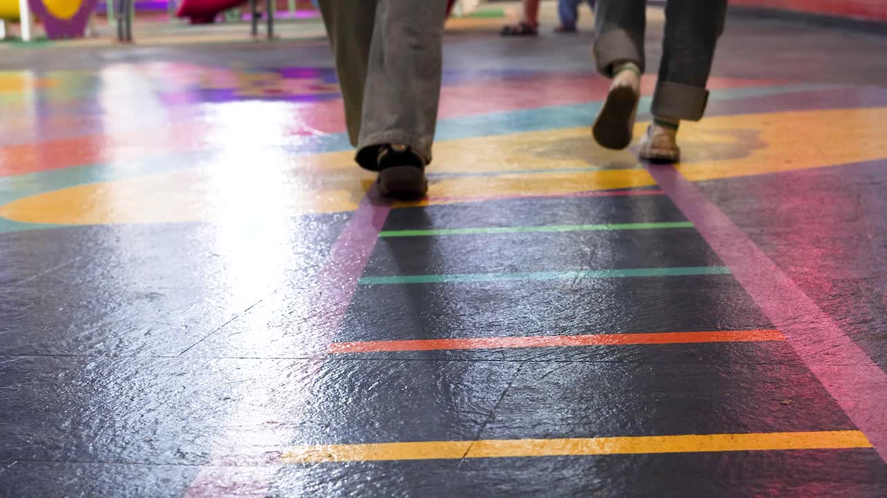 Low-angle tracking shot of two people walking on vibrant striped indoor playground with soft lighting