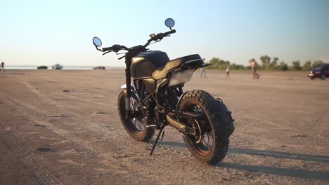 Black-bike-parking-on-the-ground-or-sand-close-to-the-water-in-sunset,-blurred-background-with-people-silhouette
