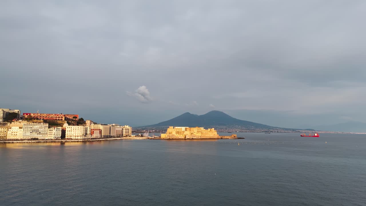 Luftaufnahme von Neapel zur goldenen Stunde mit Castel dell’Ovo, Vesuv, ruhigem Buchtgewässer und einem roten Frachtschiff am Horizont unter einem sanften Abendhimmel