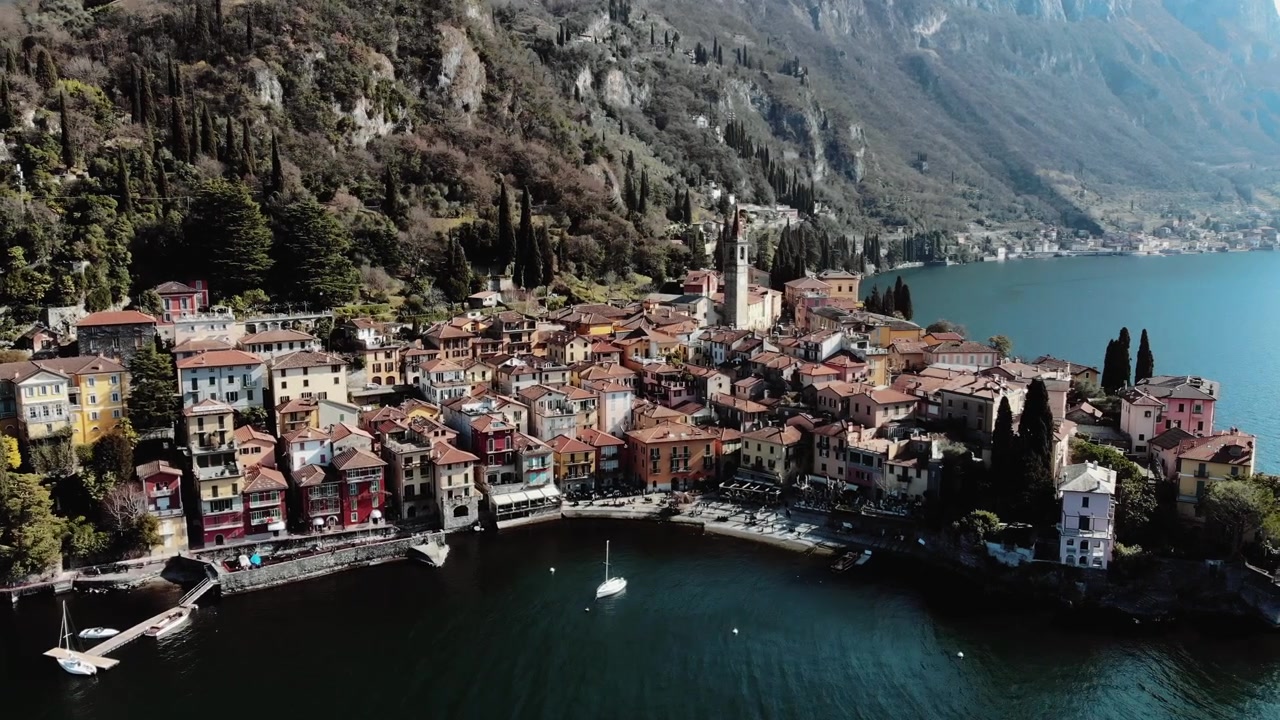 Vista aerea con drone di Varenna sul Lago di Como in Lombardia, Italia, con la Chiesa di San Giorgio sul lungolago, edifici colorati, uno sfondo montuoso e la riva del lago.