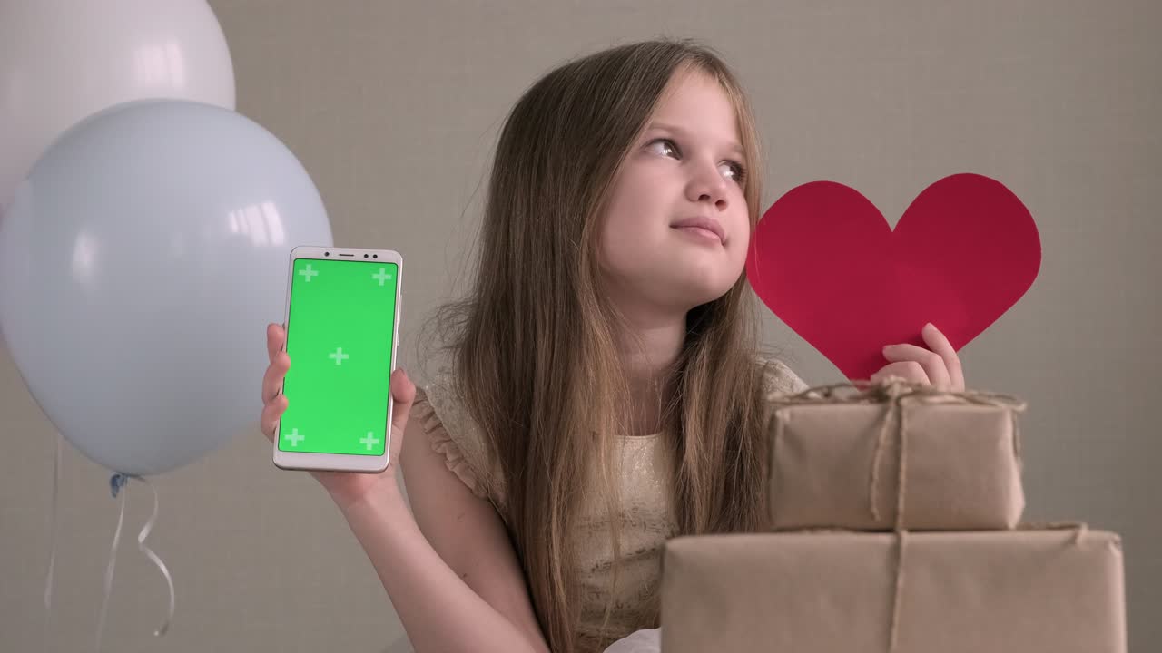 귀여운 소녀는 녹색 화면으로 드폰, 휴대전화, 전화 및 은 심장을 보여줍니다. 광고를 위해 설정 된 크로마 키 초록 화면 스마트 폰. 발렌타인 데이, 사랑, 어머니의 날