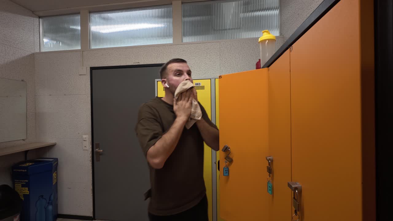 Tired man drying sweat with a towel in a gym locker room after exercise, showing post-workout exhaustion, recovery, and realistic fitness routine in a changing area