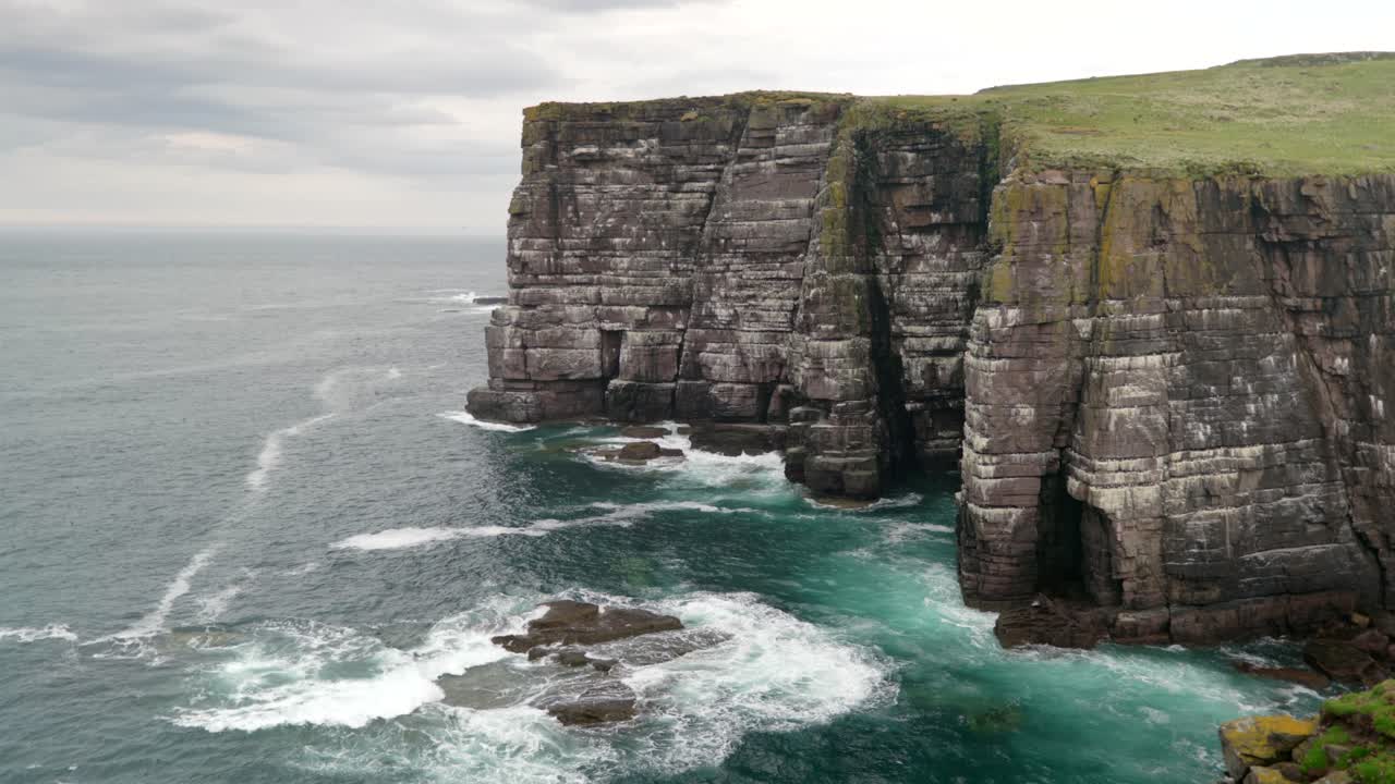 Waves slowly crash over rocks and against a tall and imposing sea cliff in the deep teal coloured ocean while seabirds fly around the cliffs of a seabird colony of guillemots on Handa Island, Scotland