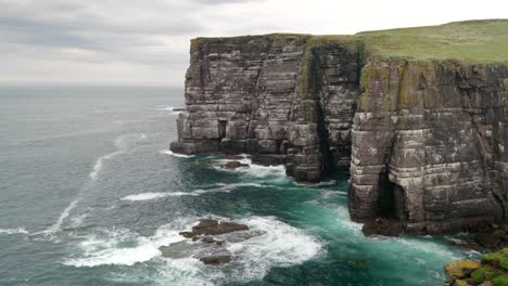 Waves-slowly-crash-over-rocks-and-against-a-tall-and-imposing-sea-cliff-in-the-deep-teal-coloured-ocean-while-seabirds-fly-around-the-cliffs-of-a-seabird-colony-of-guillemots-on-Handa-Island,-Scotland