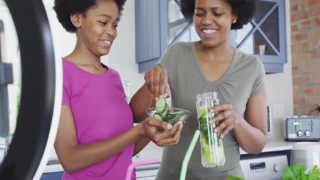 madre y hija afroamericanas felices preparando una bebida saludable, haciendo un video usando un teléfono inteligente