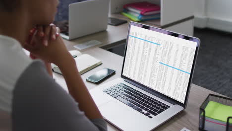 mujer biracial sentada en el escritorio viendo el procesamiento de datos de codificación en la pantalla de la computadora portátil